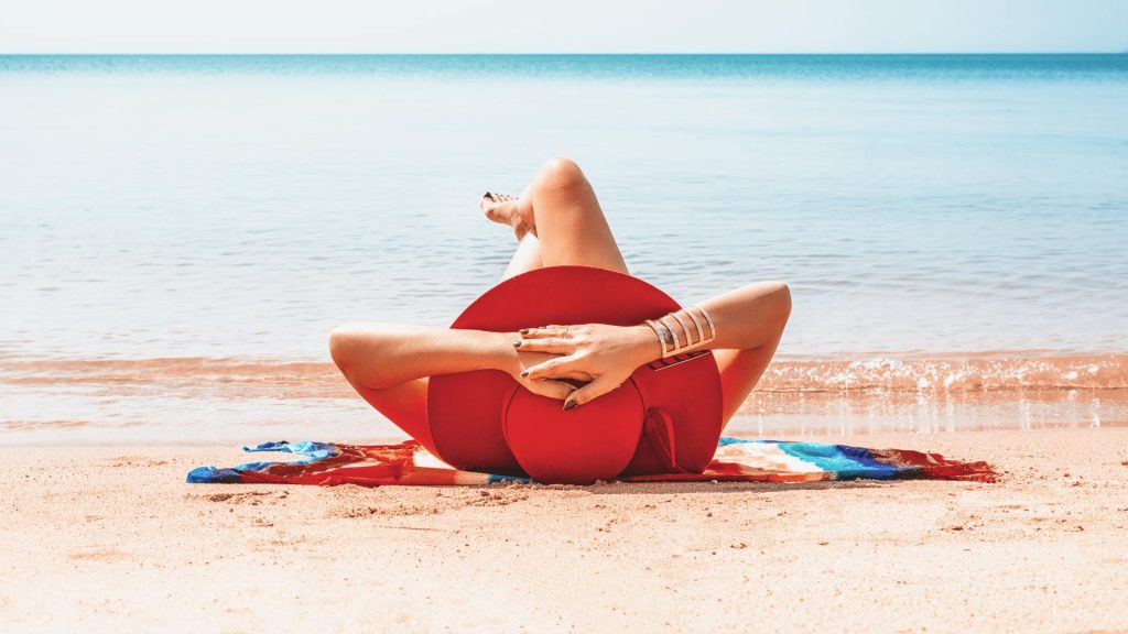 Donna con cappello rosso prendere il sole sulla spiaggia, davanti al mare